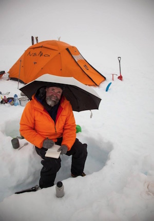 Photo of Craig Childs in deep snow under an umbrella, in front of an orange tent